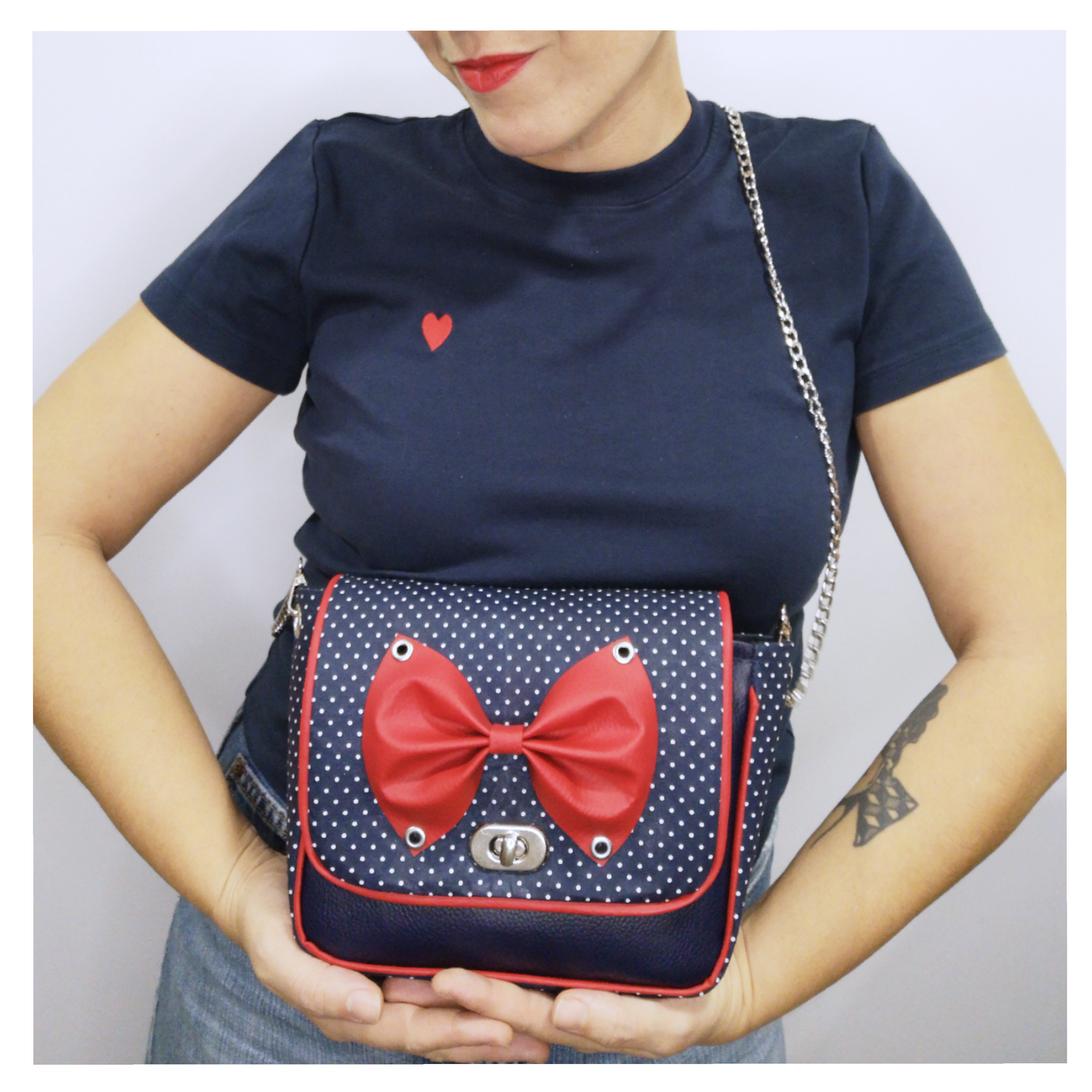 Sac à main à rabat motif bleu marine à petits pois blancs, base décorée d'un joli noeud rouge en simili cuir fixé par des oeillets argent. Muni d'un fermoir tourniquet argent et d'une chaîne argent bandoulière amovible.