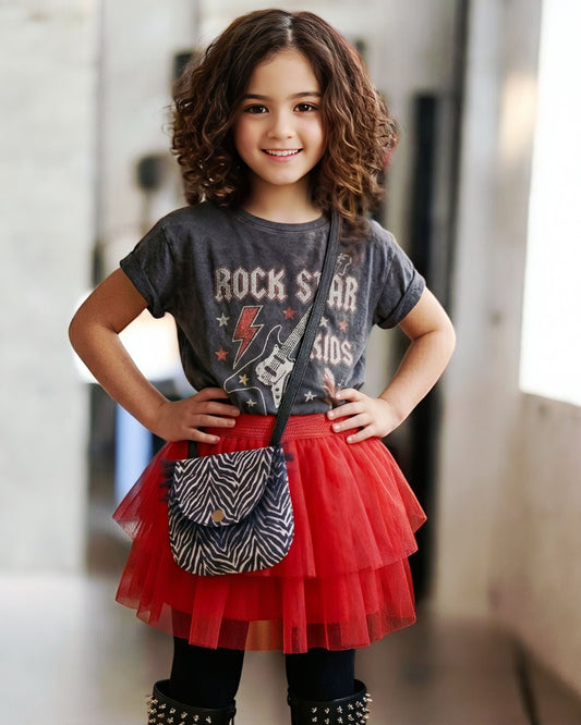Petite fille au look rock portant un t-shirt "Rock Star Kids", une jupe tutu rouge, des bottes cloutées et le sac bandoulière enfant Bonnie Zebra avec tulle noir.